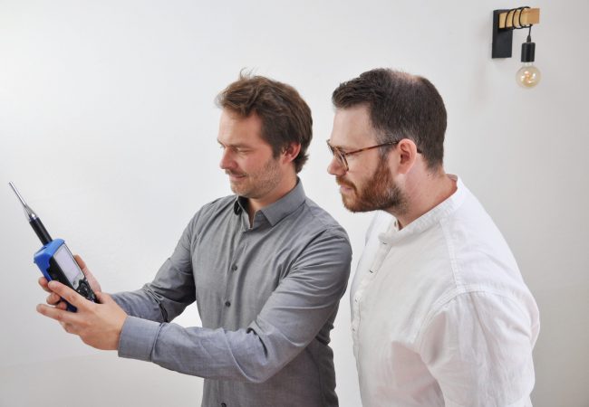 Hier sehen Sie ein Bild der beiden Gründer Daniel und Sebastian von akustik.inklusiv. Daniel hält ein Messgerät in der Hand und beide schauen darauf.